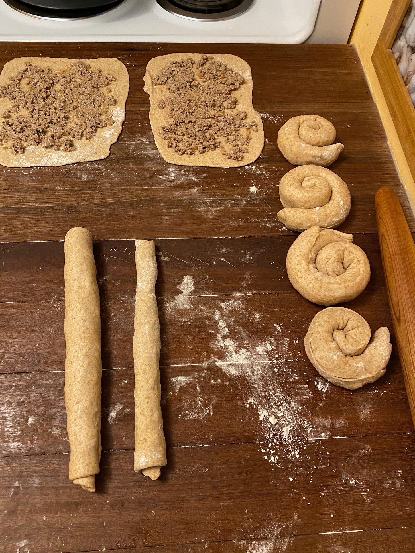 Flatbreads in various stages on a kitchen counter: two open rectangles spread with filling, two long ropes of rolled up dough, and four spiral coils.