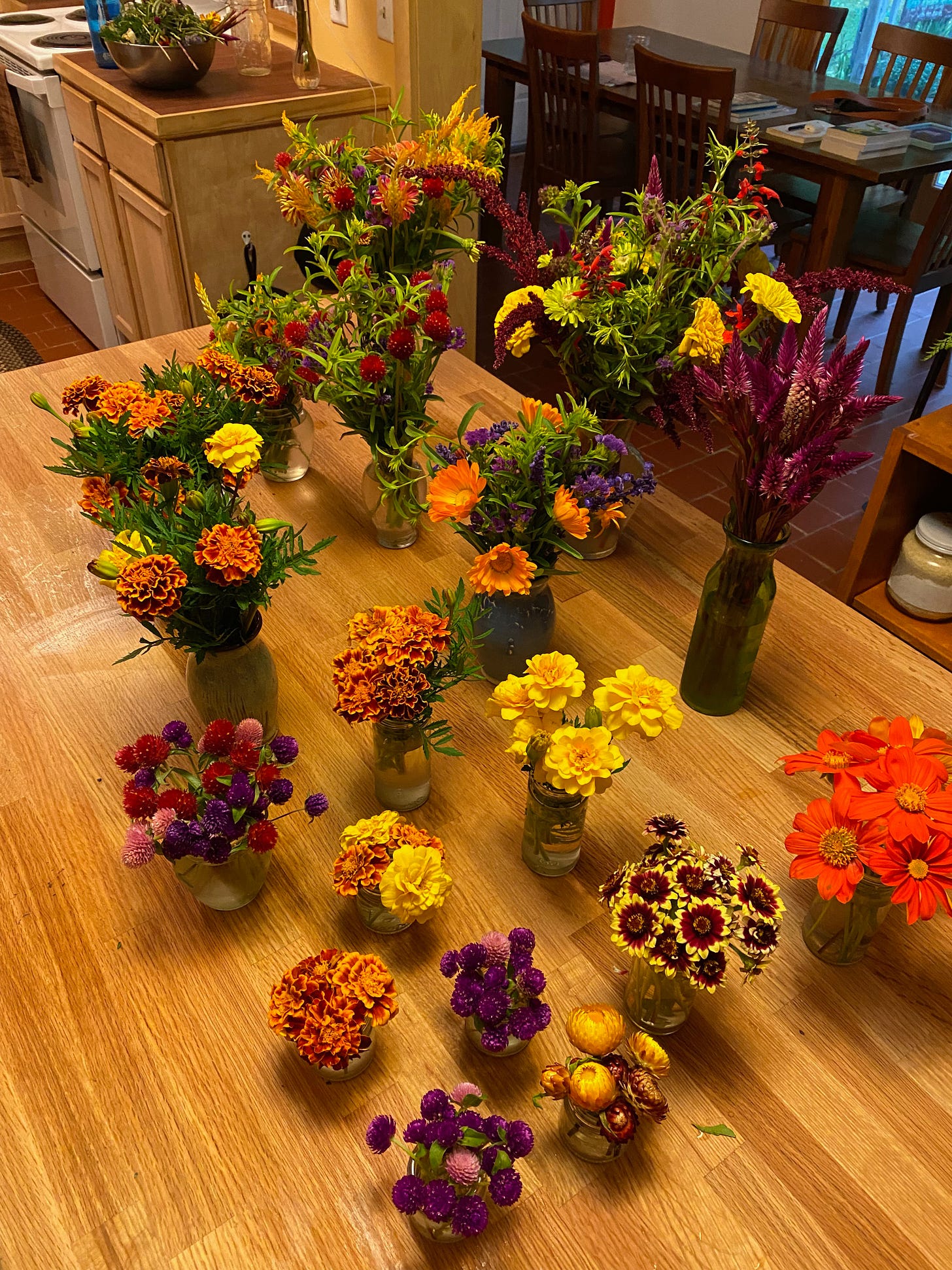 View of a kitchen island filled with vases of flowers. There are several small glass jars of yellow and orange marigolds, pink and red gomphrena, Mexican sunflowers, and zinnias. There are also several larger vases with mixed bouquets, mostly in shades of red, yellow, orange, purple, and pink.