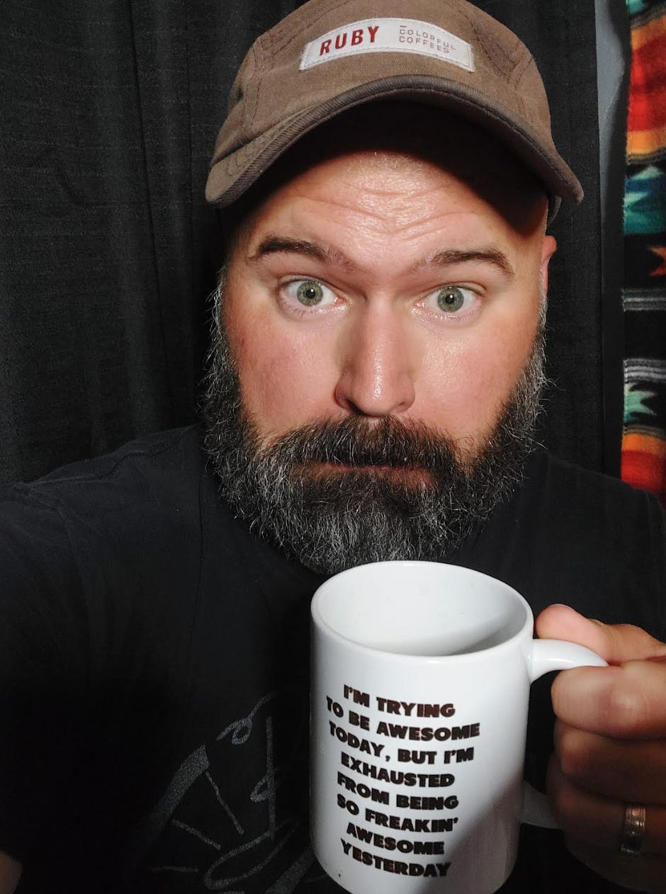 A selfie of a white man with a lush salt and pepper beard, green eyes and a gray baseball cap holding a white coffee cup up to the camera and making a faux panicked face.