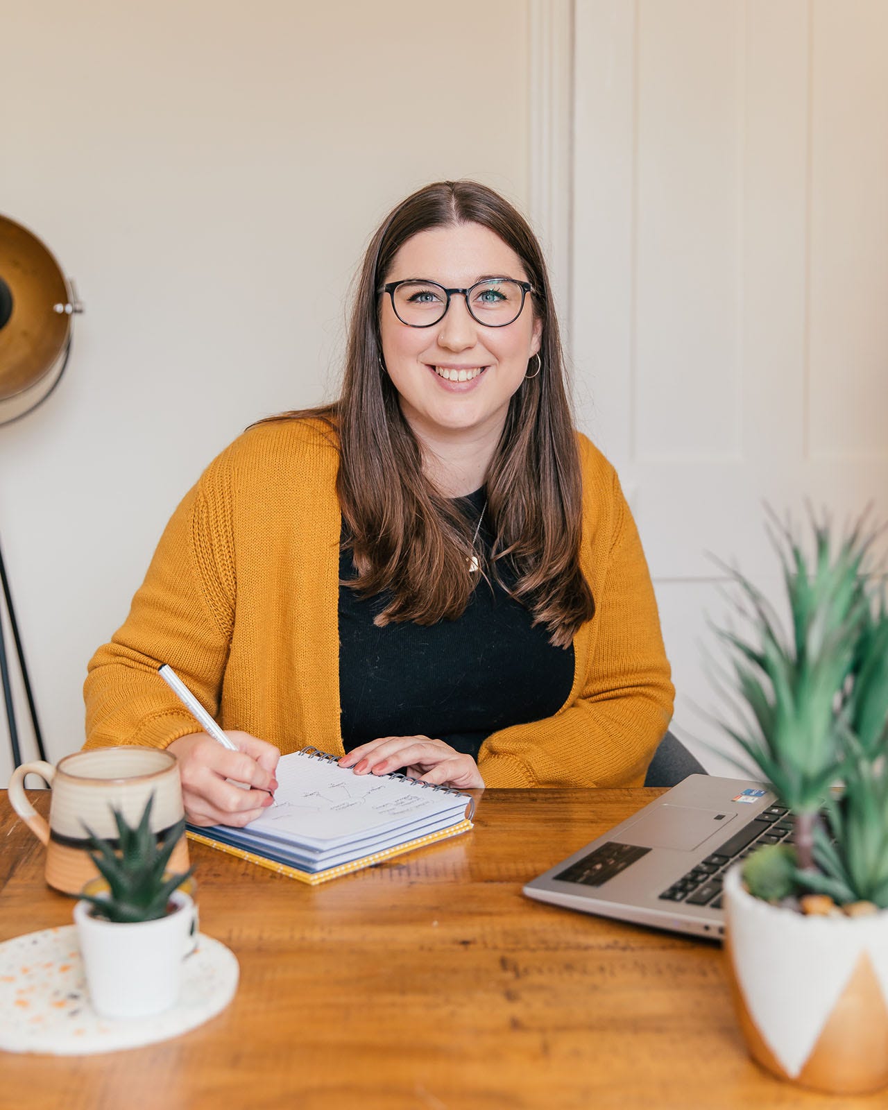 Rachel Baker, freelance copywriter and founder of The Ethical Copywriter, sits at her desk and maps out a content strategy in her notebook. On her desk is a laptop, plants, candle and a mug. She wears an orange cardigan, is gazing straight ahead at the camera and is smiling.