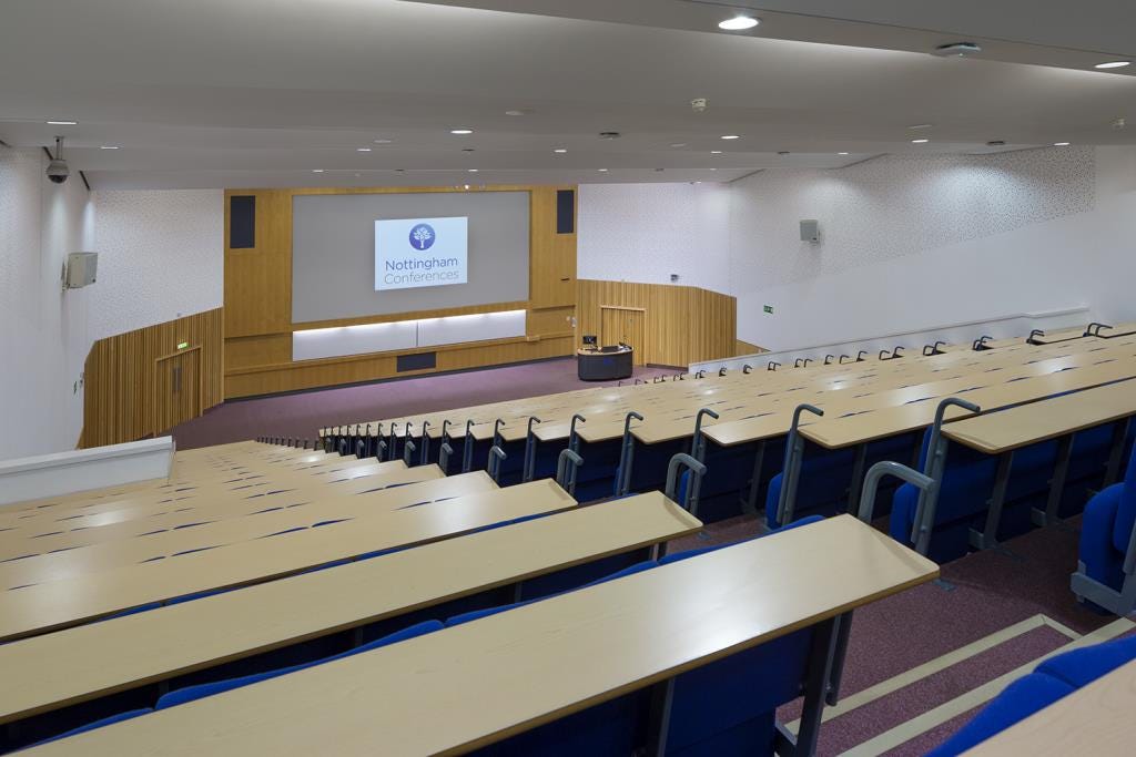 An empty tiered lecture hall. The photo is taken from the back row of the lecture hall, looking toward a stage with a large screen.