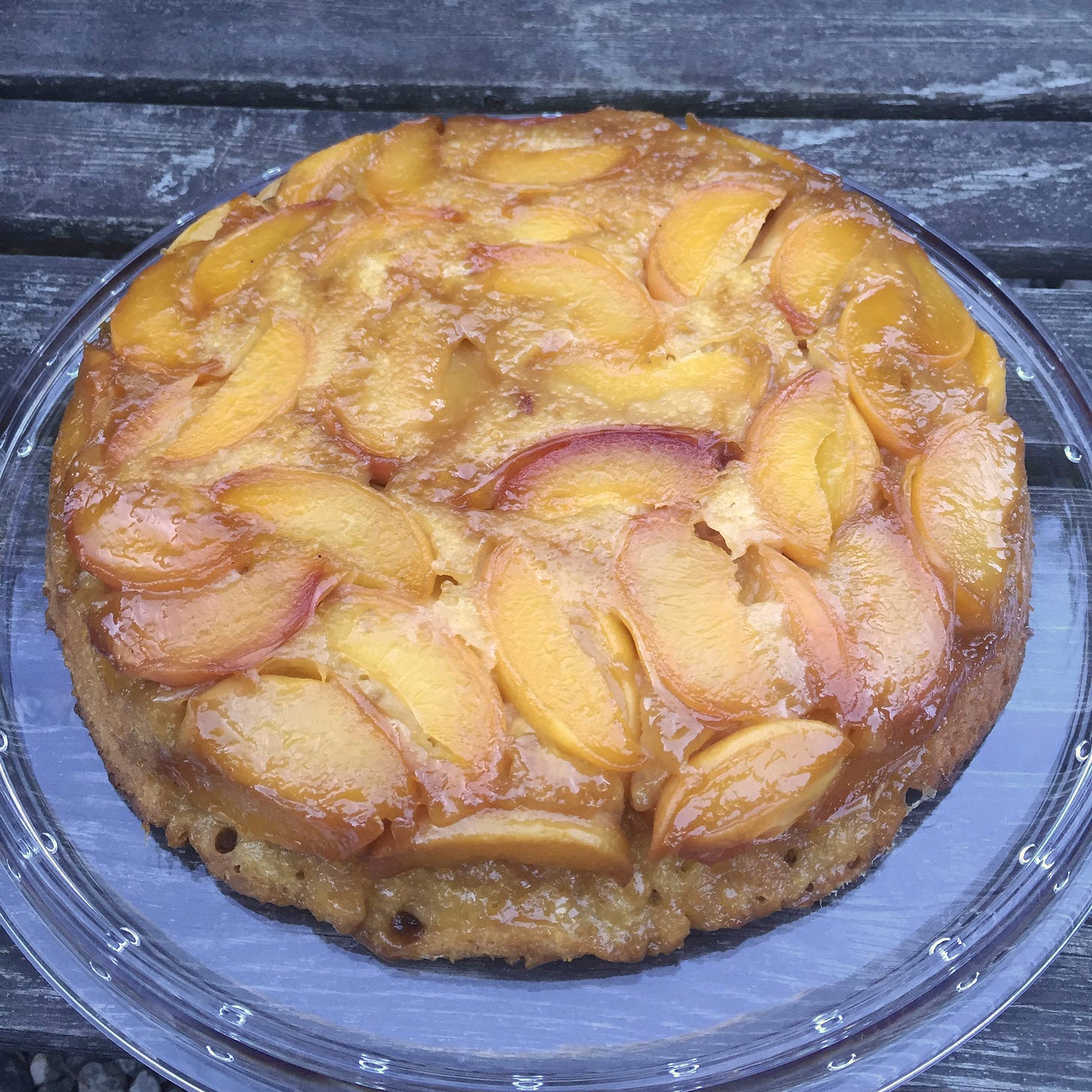 On a clear cake plate, an upside-down cake with yellow and reddish peach slices, caramelized at the edges.