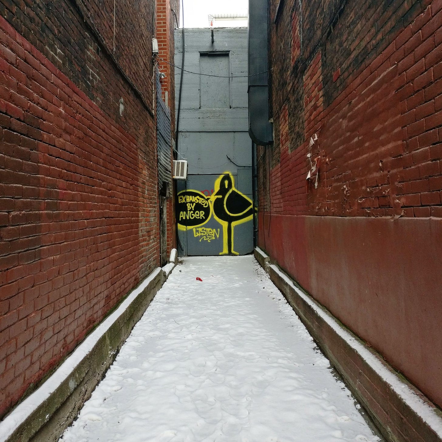 A dead end alley in Montreal with a graffit bird at the end, saying "exhausted by anger." The bird is black with a yellow outline on a narrow gray wall. The other two walls are red brick, patched and painted. The ground is covered in snow.