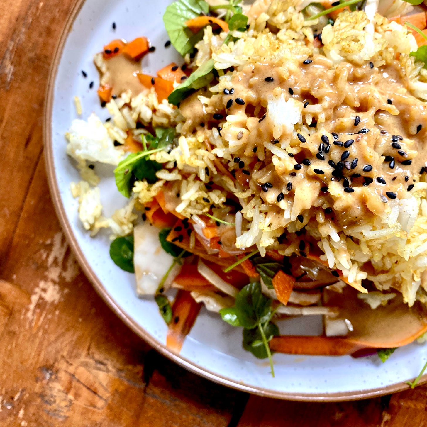 overhead shot of a plate topped with salad consisting of carrots, white cabbage, crispy rice, black sesame seeds, salad leaves and peanut dressing. Plate is a pale colour with a brown edge, placed on top of a wooded table