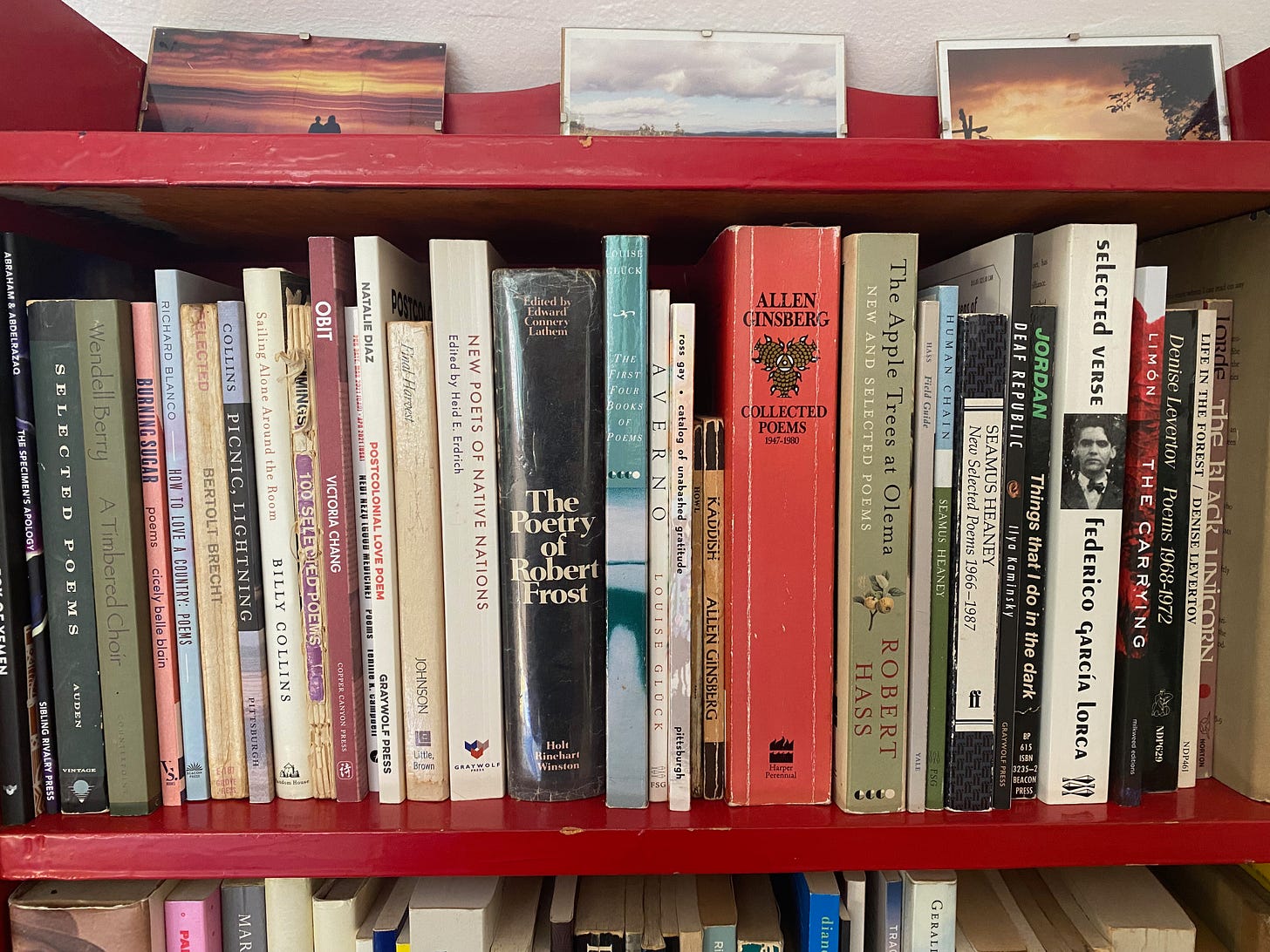 A closeup of a shelf of poetry books, including several large volumes of Robert Frost, Allen Ginsberg, and Robert Hass, as well as some slimmer, newer collections. Three photos of sunsets and the sky sit on top of the shelf.
