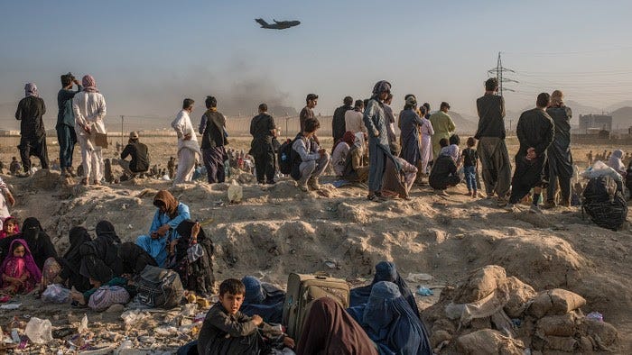 Women and children sit on dusty rocks with men standing behind, facing distant airport walls and watching a military plane taking off