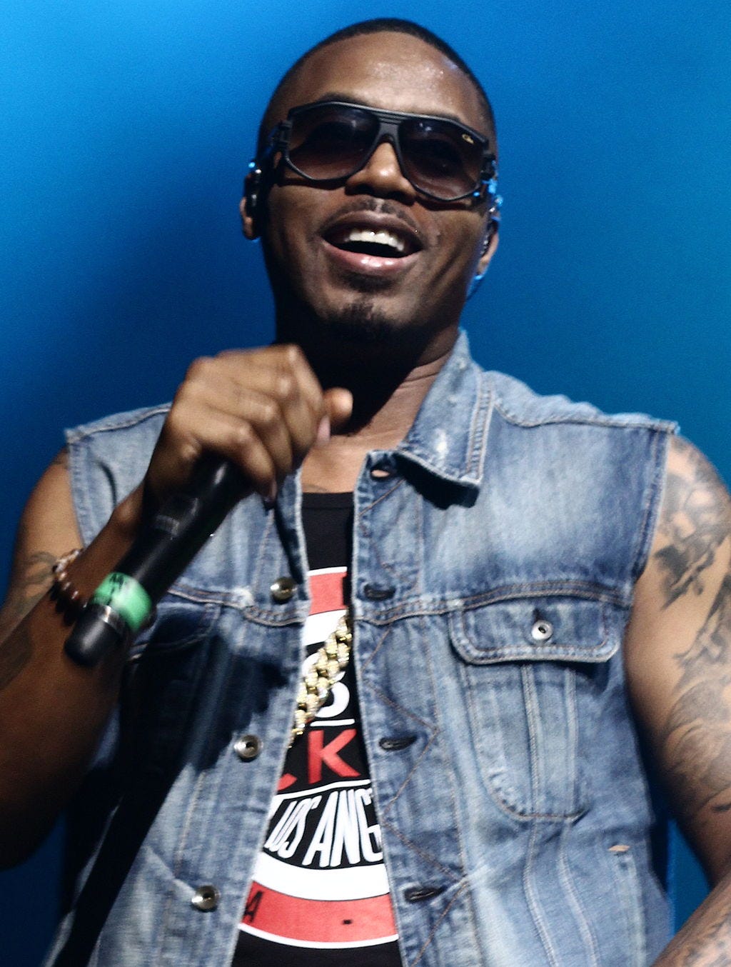 A black man with sunglasses and a sleeveless denim jacket, smiling with a microphone in hand