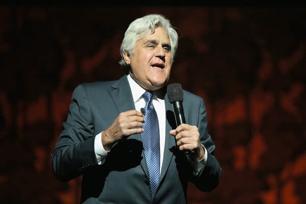 Jay Leno, the comedian and TV host, speaks into the mic while onstage. He is wearing a dark charcoal jacket and blue tie.