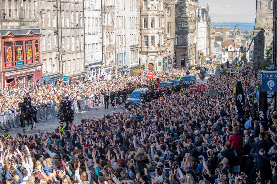 A large crowd stands on either side of a street as a procession of cars follows a hearse carrying Queen Elizabeth's coffin.