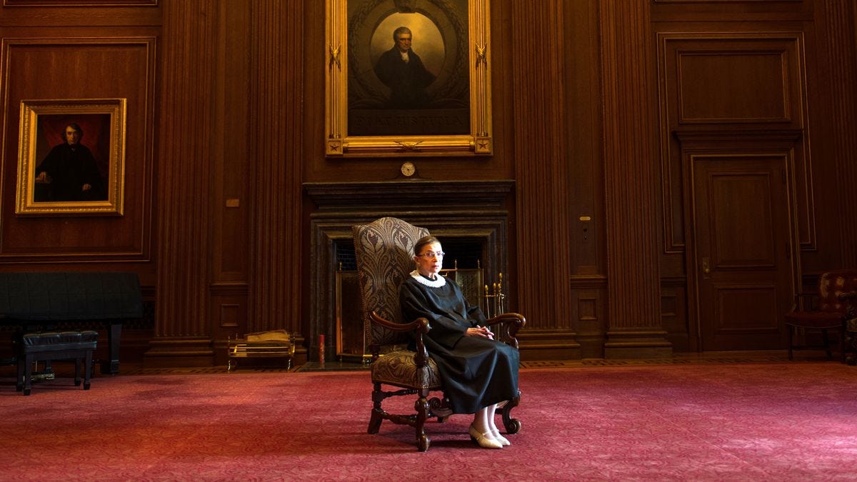 Supreme Court Justice Ruth Bader Ginsburg sitting in a high-backed chair in front of a fireplace and below a large portrait.