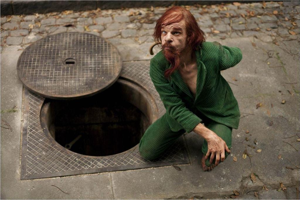 A film still from Holy Motors showing Mr. Oscar in a green suit crouched beside an open manhole cover.