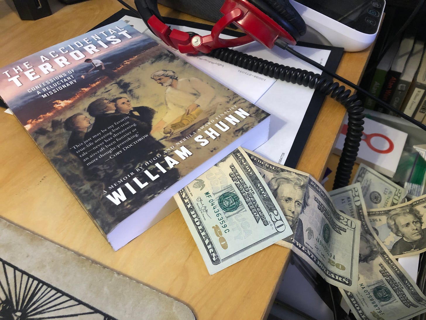 A copy of a paperback book, The Accidental Terrorist by William Shunn, lies on a cluttered desk with twenty-dollar bills spilling out of it.