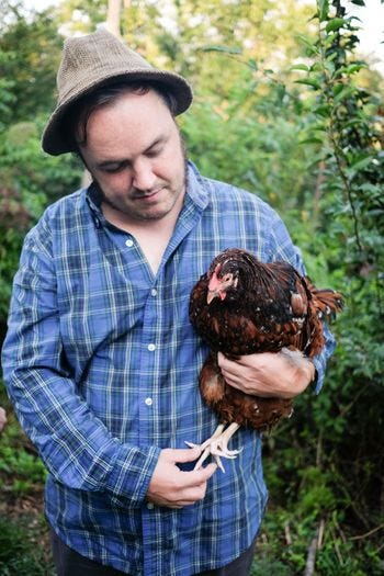 A white man wearing a blue flannel shirt and a brown-grey fedora is standing here in a forest background, with one branch prominently in the foreground. The guy, who is Erik Simpson, is holding a dark brown speckled chicken, and looking toward it instead of the camera.