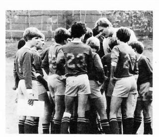 A black and white image of the soccer team in a huddle: boys in long sleeve jerseys and shorts and knee socks. Todd's back is to the camera, and Brian is looking sideways, notebook in hand.