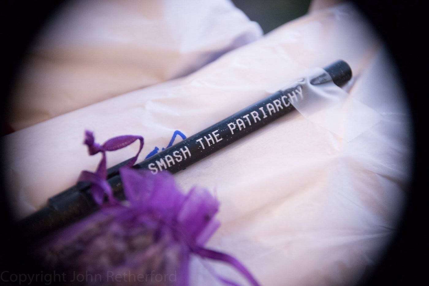 A picture of a black pen that has "SMASH THE PATRIARCHY" written on the side in white font in capital letters. The pen is laying on white tissue paper. There is a purple ribbon herb pouch in the bottom left of the picture.