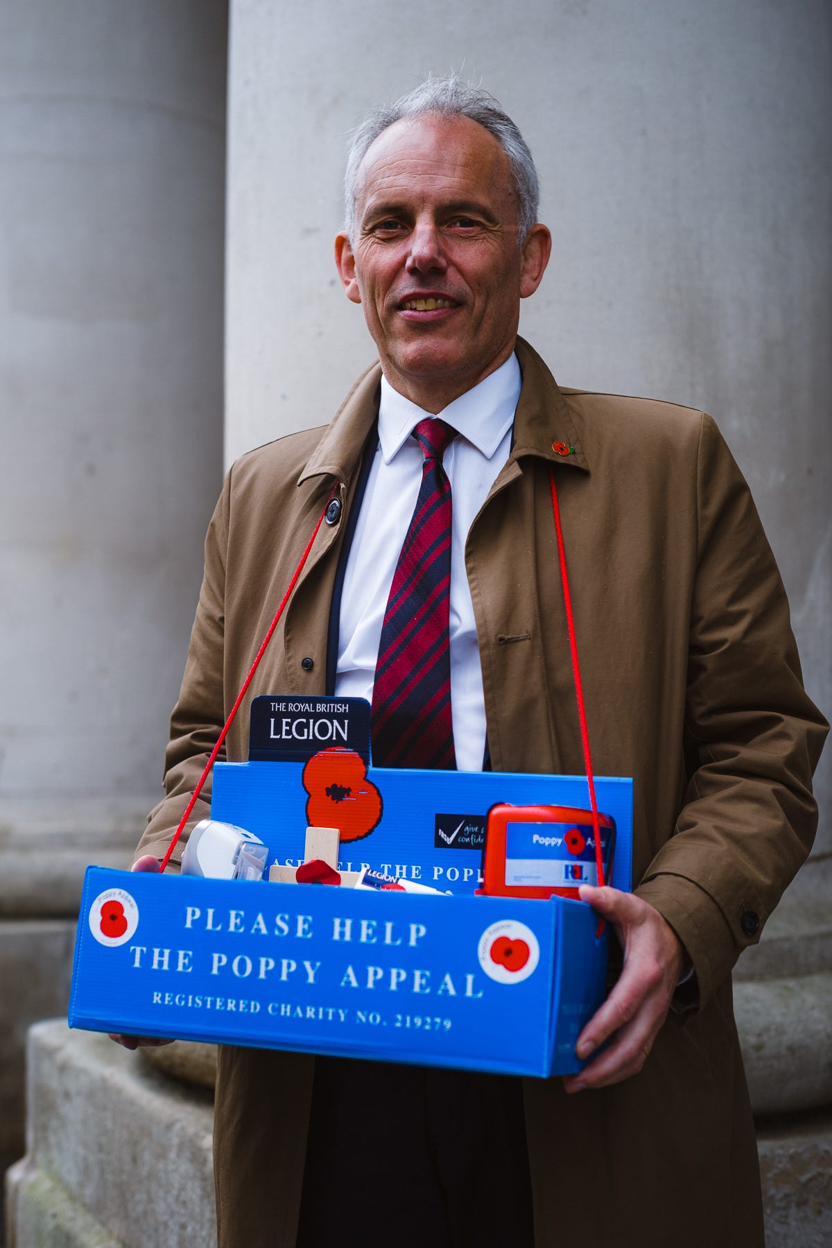 Portrait of a caucasian man with gray hair in a white shirt, red and navy tie and tan trenchcoat asking for donations for the veterans poppy appeal