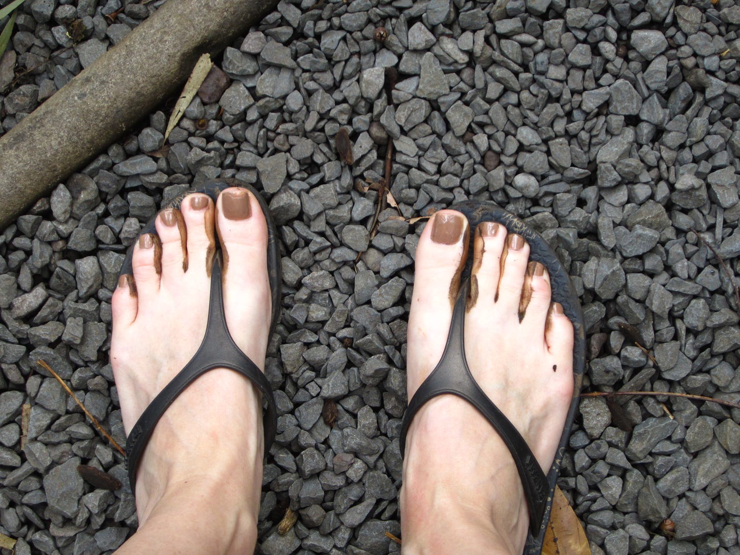pair of bare feet in flip-flops, standing on grey crushed stones, brown painted toenails, brown Hawaiian mud squishing up between toes.