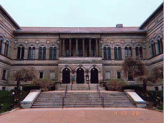 Carnegie library, made of tan stone. A set of stairs leads up to three doors under archways, and the building's two arms extend toward the viewer.