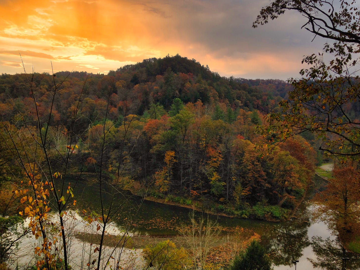 The orange, red, brown, and green of the forest trees overlooking a small mountain lake with the setting sun on the left