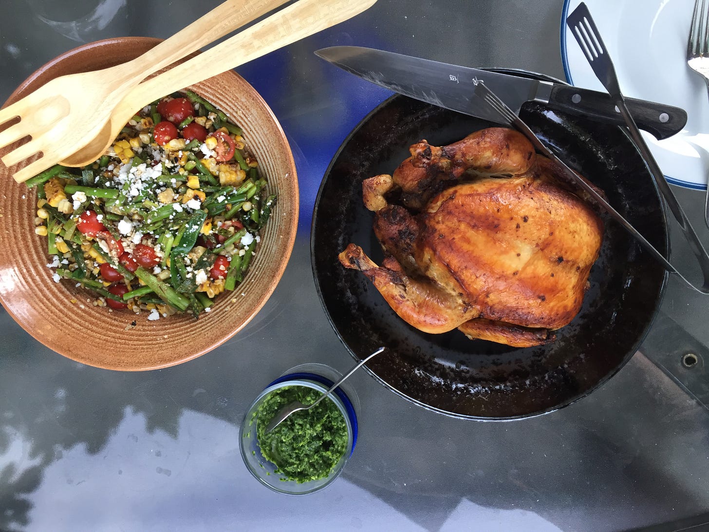 From above, an outdoor table with a bowl of corn, asparagus, and tomato salad sprinkled with feta. A wooden fork and spoon sit at the edge of the bowl. To its right, a black dish with a darkly browned roast chicken. A large knife and a pair of tongs rest on the edge of the dish. Between the two dishes is a small glass bowl of chimichurri.