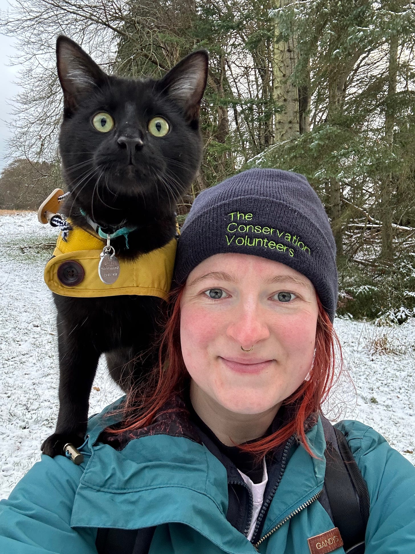 Rosie is wearing a blue coat and a blue beanie, with a black cat (Jasper) stood on her shoulder. Jasper is wearing a yellow coat. They're stood in front of a group of trees, there's snow on the ground.