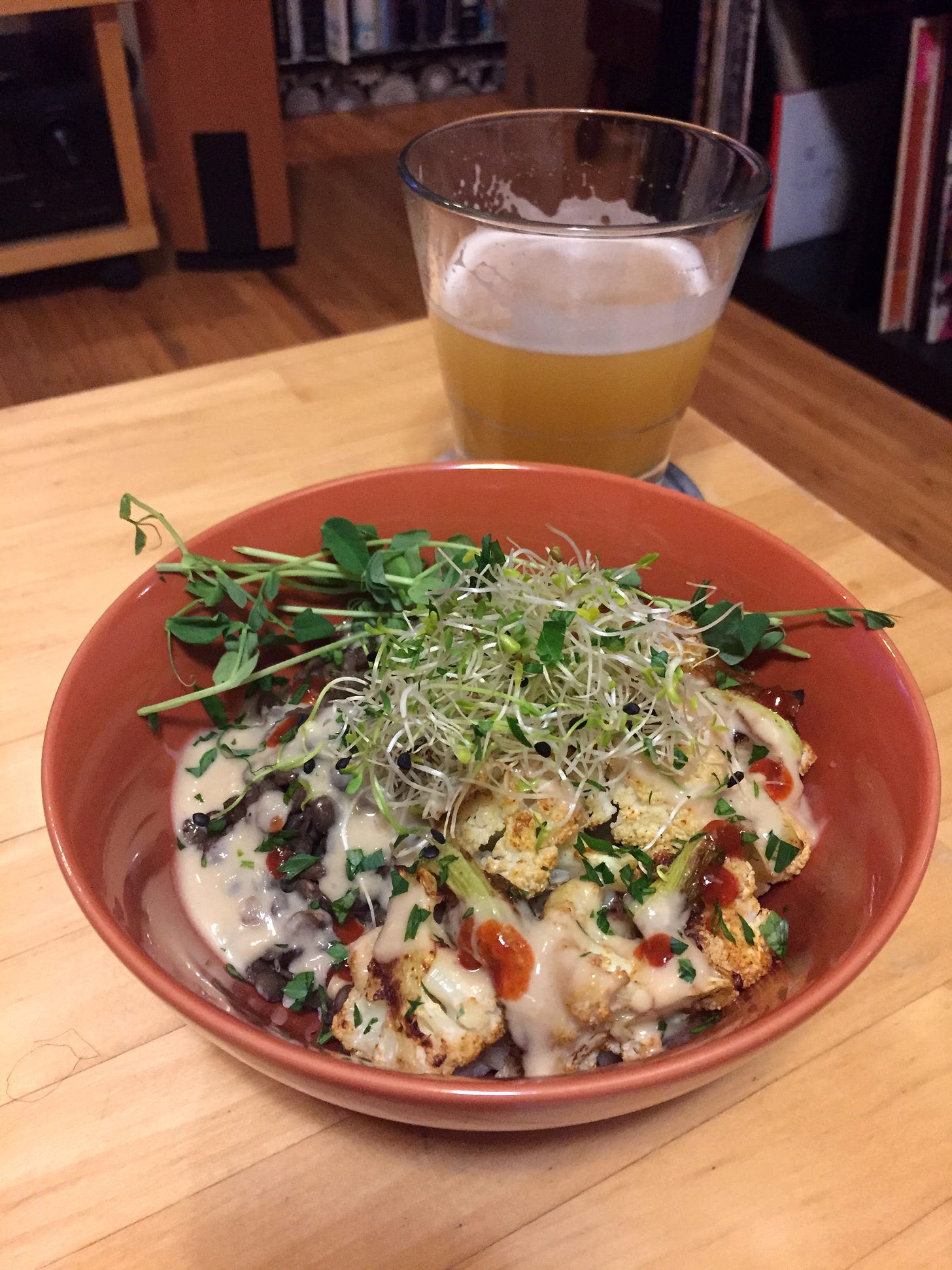 An orange bowl full of rice and lentils with roasted cauliflower and lots of tahini sauce, with a pile of sprouts and pea shoots on top. Red spots of sriracha dot the sauce in places.