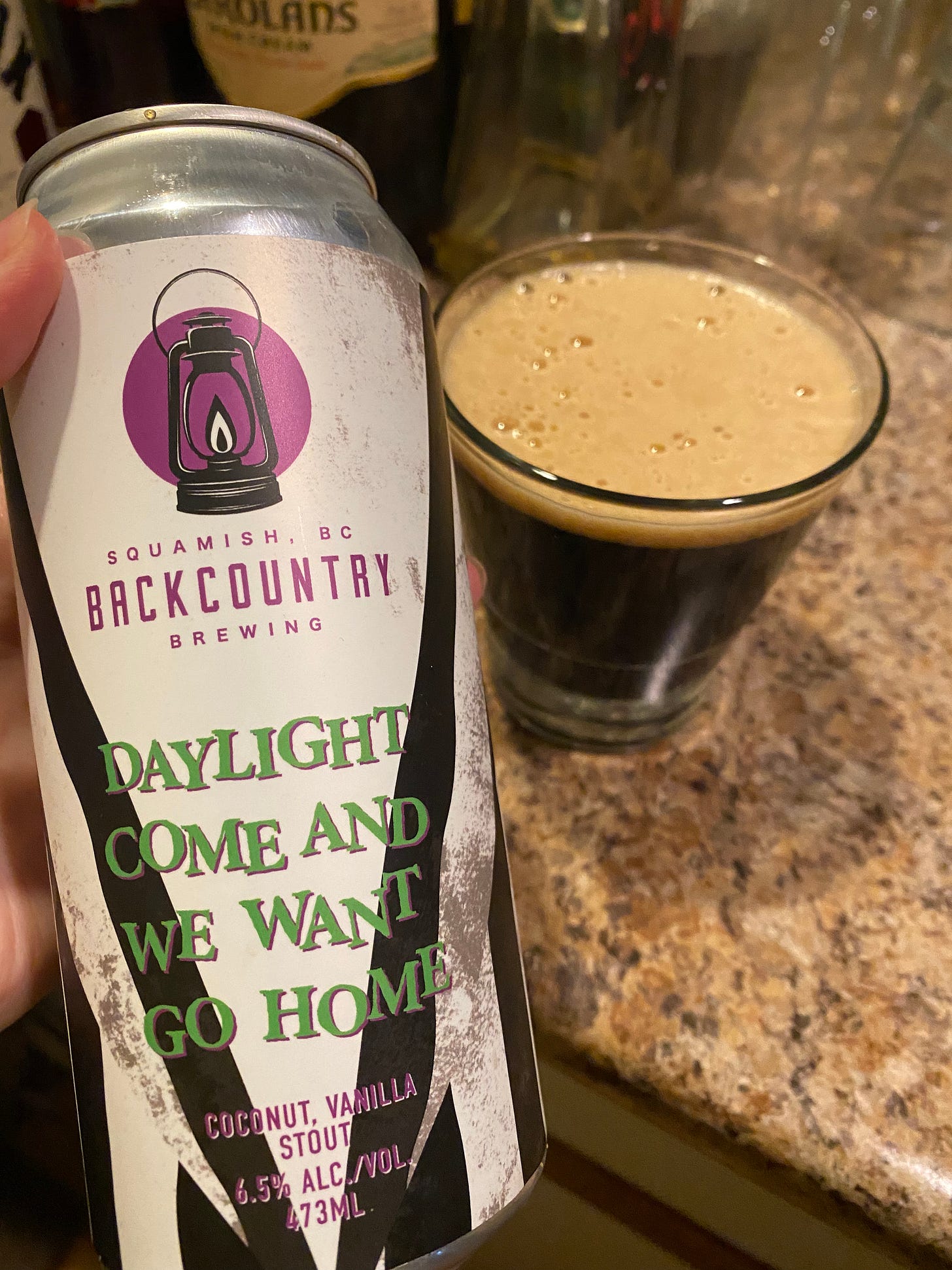 A white and black can of beer with purple and green text in the style of "Beetlejuice", reading "Daylight come and we want go home". In the background behind the can is a glass of dark beer with a light brown foam on top.