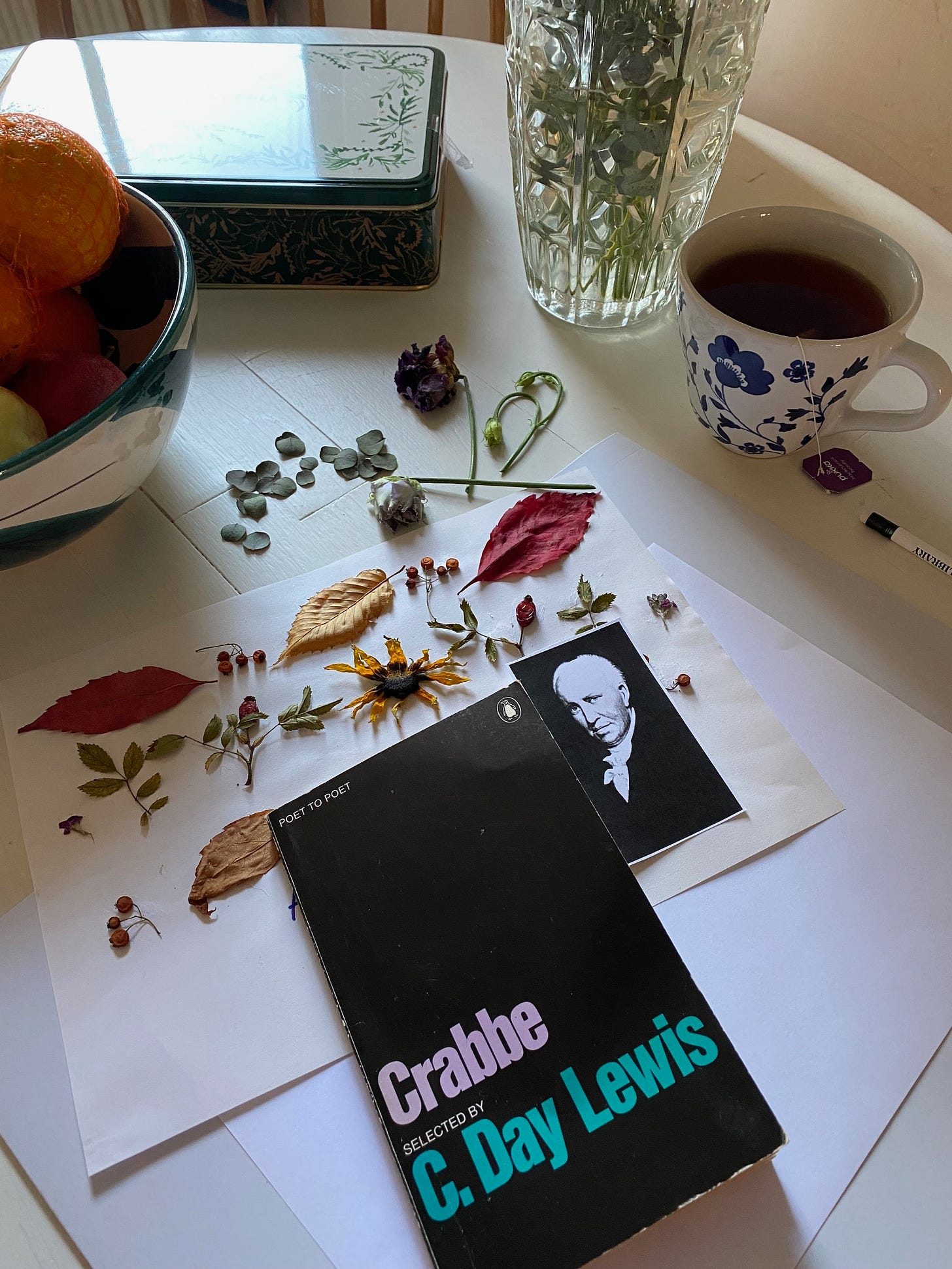 A cluttered table on which can be seen a book of poems by George Crabbe, dried flowers arranged on a page and a cup of tea.
