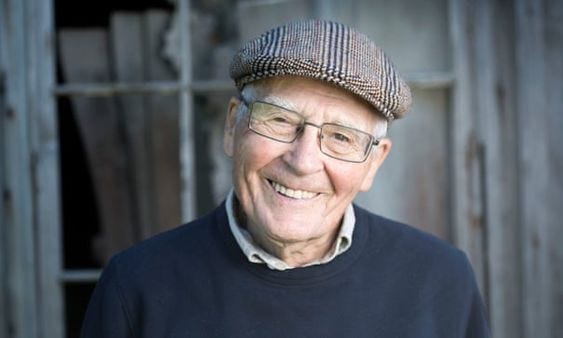 Foto de um idoso branco de boina e óculos, bem simpático, sorrindo com os dentes à mostra, com a cabeça levemente inclinada para a direita.