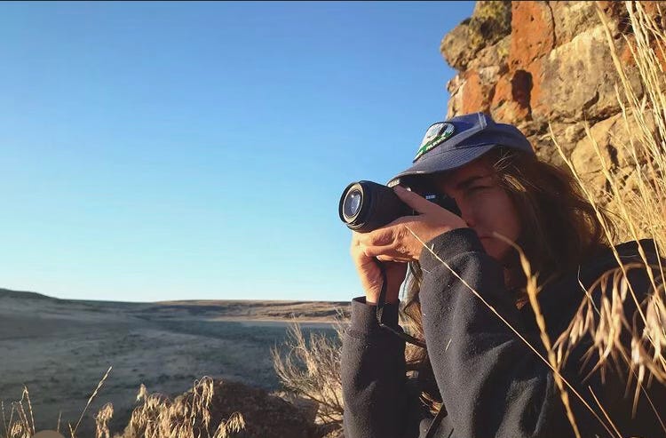 Shelby Herbert stands in front of a cliff face surrounded by tall grass. She is pointing a camera at something out of frame.  