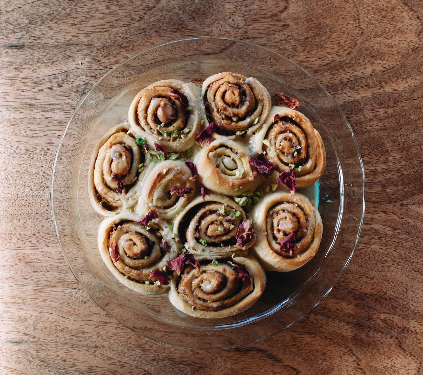 ten rose and cardamom buns arranged in a clear pie dish sitting on a table top. the photo is taken from above looking down on them. they are topped with dried pink rose petals and chopped pistachios and drizzled with glaze. 