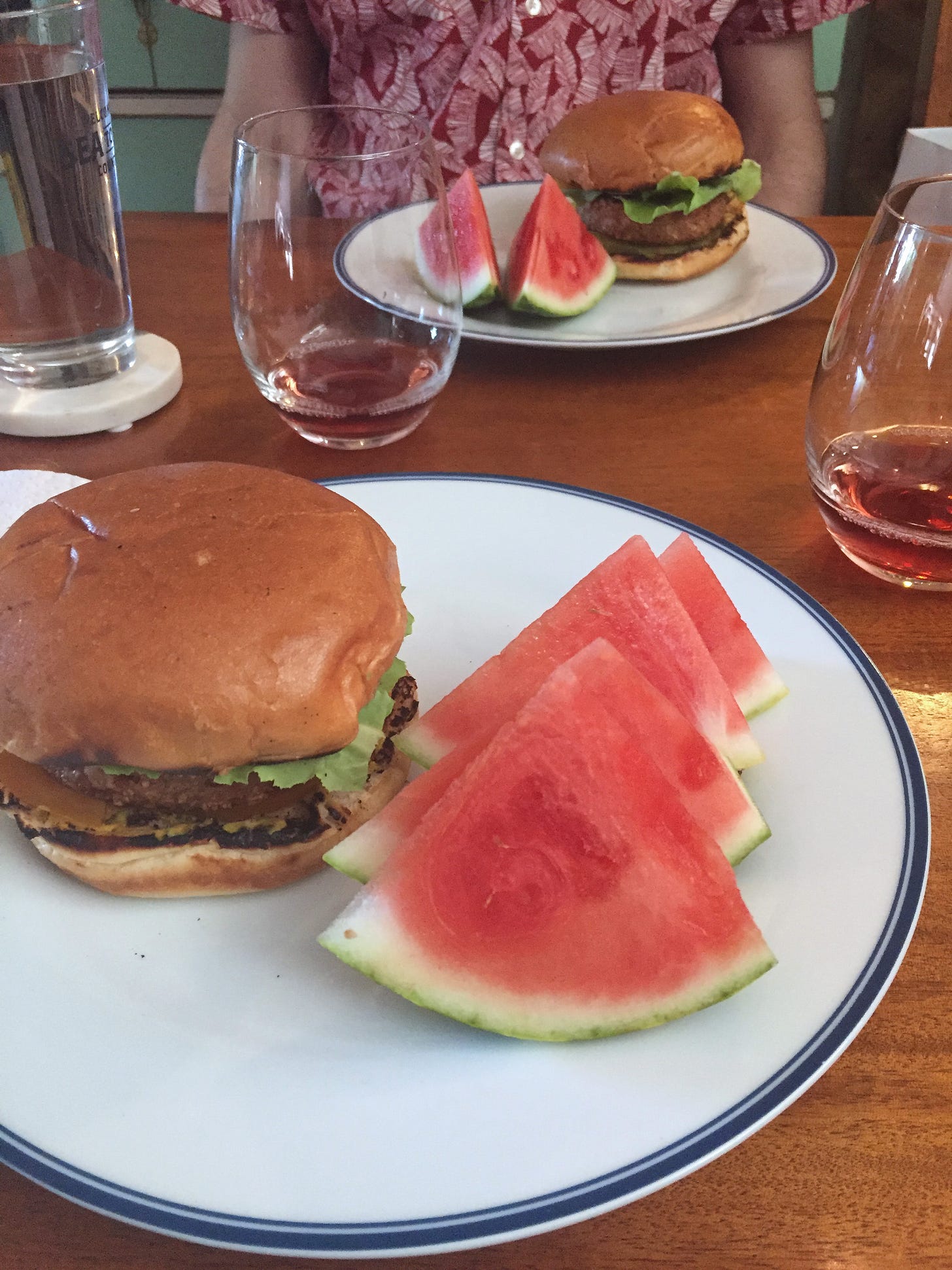 two plates with slices of watermelon and veggie burgers on brioche buns. small glasses of rosé sit at the top corners of each plate.