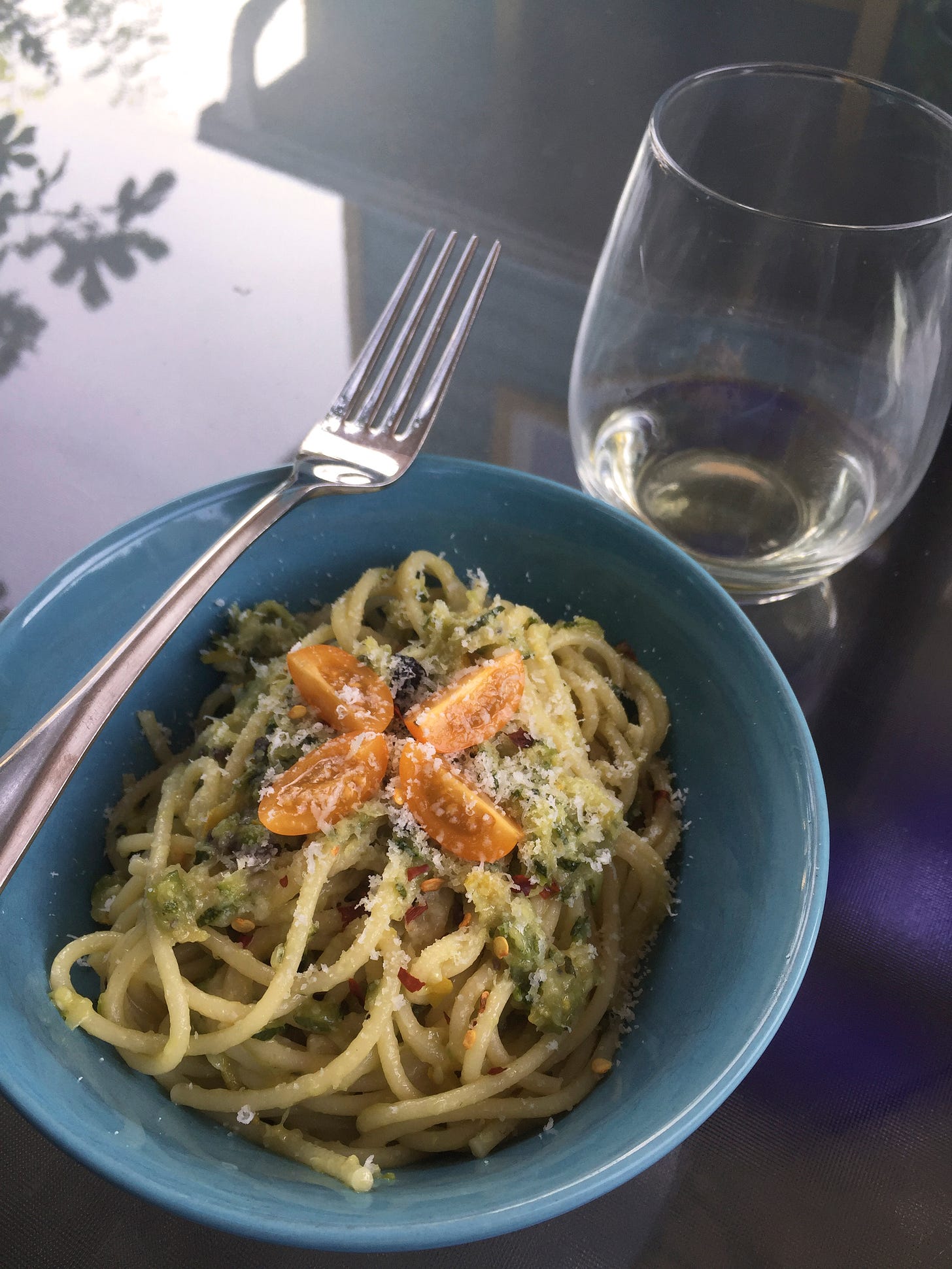 A blue bowl of spaghetti in a zucchini sauce, with parmesan and a quartered orange grape tomato on top. A glass of white wine is in the background, and a fork rests at the edge of the bowl.