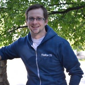 Foto de David Walsh, em meio corpo, de óculos, vestindo uma blusa em que está escrito Firefox OS, com uma árvore ao fundo.
