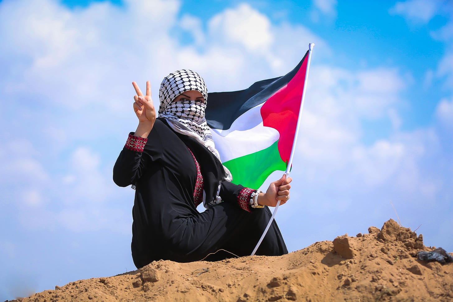 A woman wears all black and a keffiyeh as a head covering. She is facing the camera, sitting on a red dirt mound, and looking toward the camera while holding a peace sign with her left hand and a Palestinian flag with her right hand. The background is a blue sky with big fluffy white clouds.