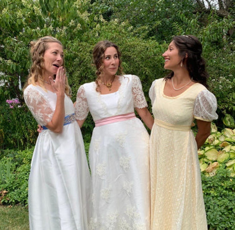 Three young women stand in a garden wearing Jennyvi Dizon designed, Regency-inspired gowns. Two are white and one is pale yellow, and all have lace sleeves and wide sashes. They are laughing and smiling together, inspired by the Dashwood sisters, Marianne and Elinor Dashwood, from Jane Austen's novel 'Sense and Sensibility.'