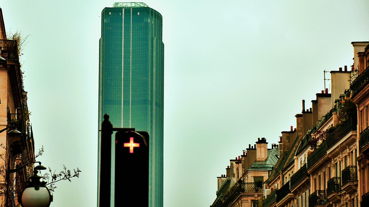 Foreground: red traffic light with "plus" symbol; background: black skyscraper against grey skies; sides: Haussmannian architecture