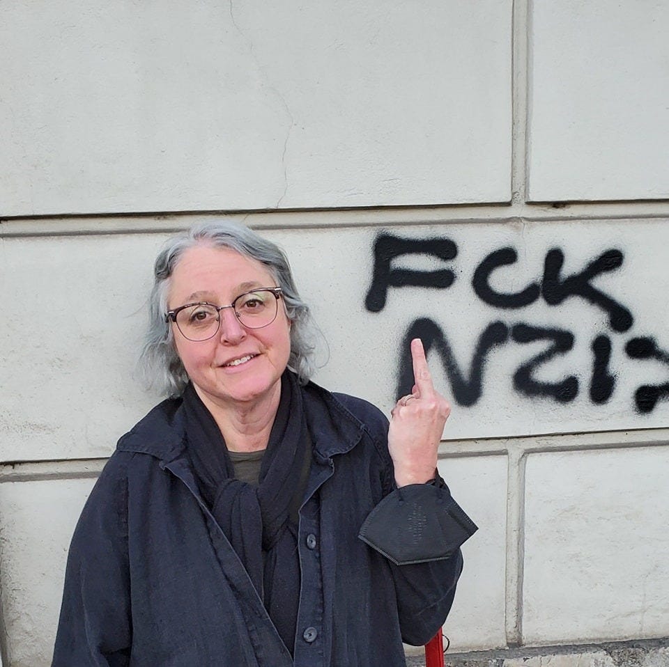 Gray haired white woman raises a middle finger in front of graffiti that reads "FCK NZIS"