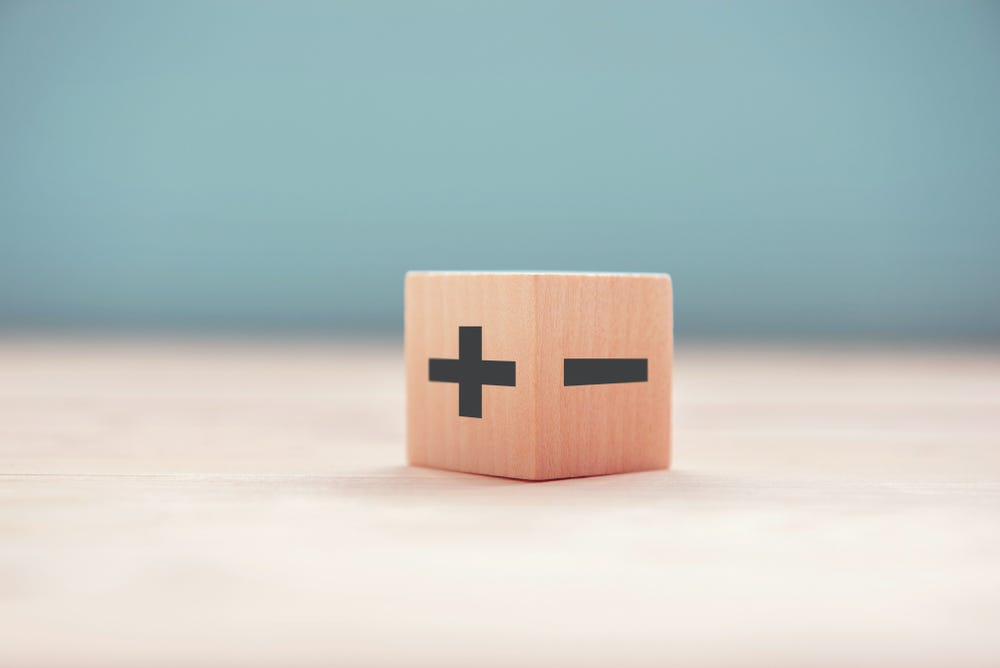 wooden die with a plus and minus symbol on the two visible sides. The background is blurred out, cream ground, blue wall