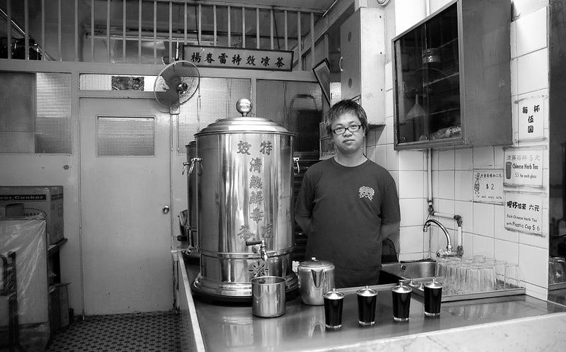 a black and white photo of a man next to a large urn of herbal tea in a Hong Kong herbal tea shop