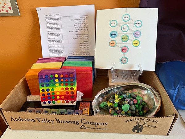 A still image of a custom omer counting kit with prayer beads, tree of life illustration, and a stack of eight canvases, two still in progress. Also a sheet of blessings for counting the omer. 