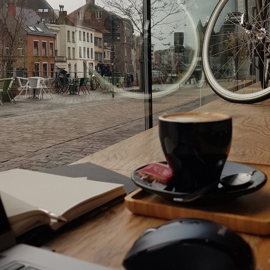 Close up of a black coffee cup and saucer on a wooden board next to an open notebook and pen, on the windowsill looking out over a cobbled street and metal tables and chairs. A bicycle is decorating the window.