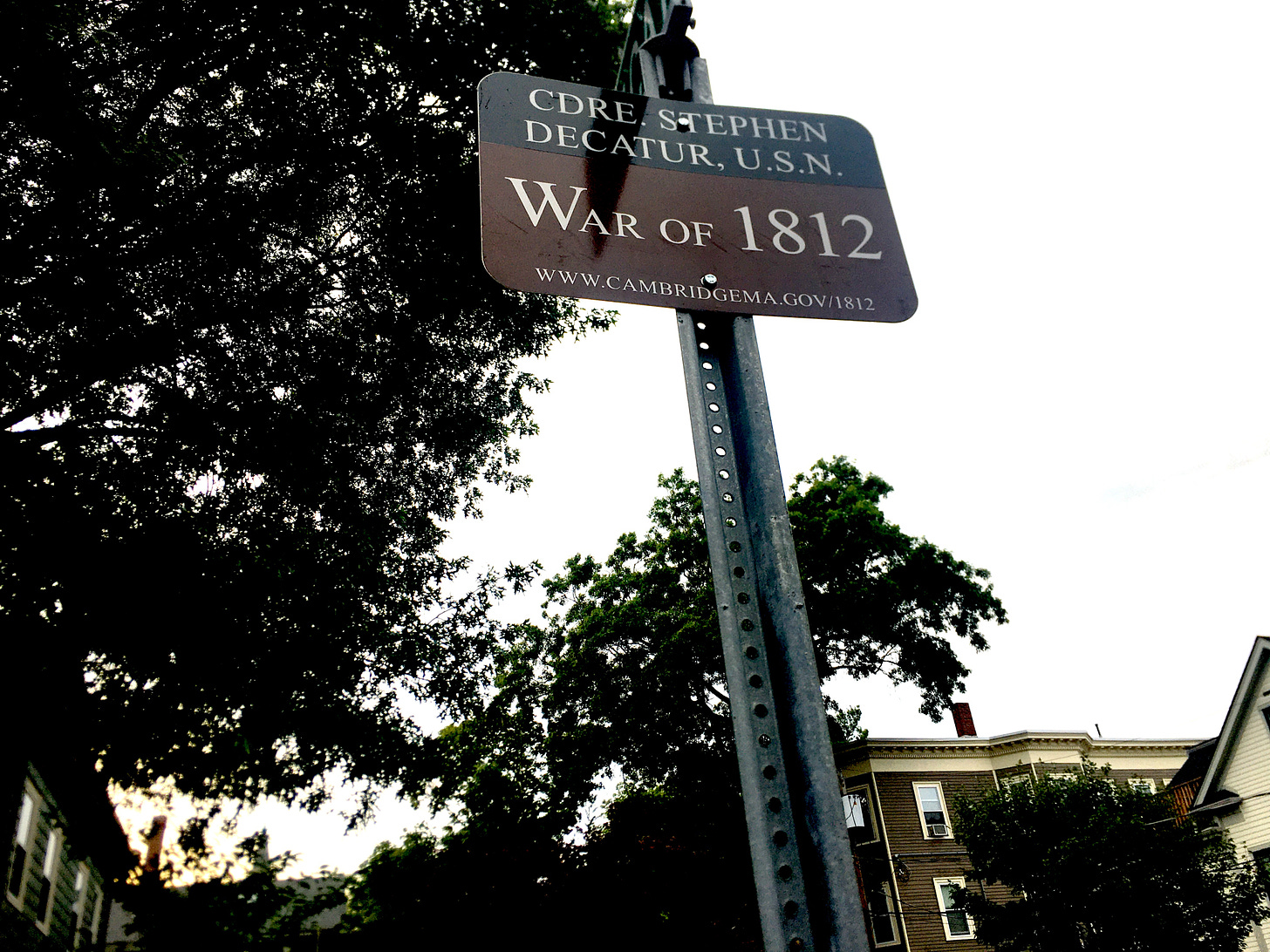 A small black and brown street sign on a corner in Cambridge reads in memoriam of a soldier, Stephen Decatur, who died in the War of 1812.