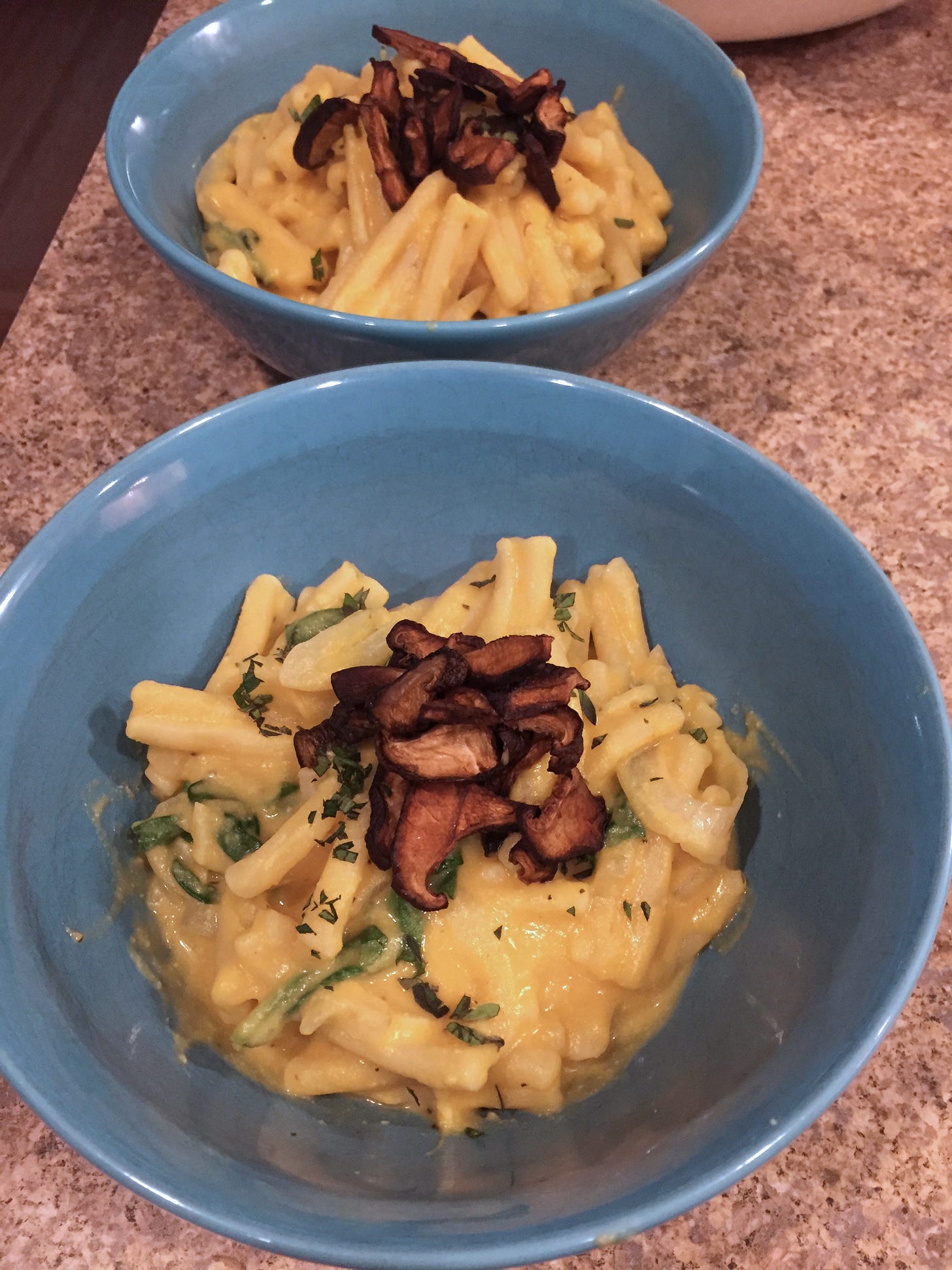 Two blue bowls with casarecce in a creamy light orange sauce, with chopped oregano and shiitake bacon on top.