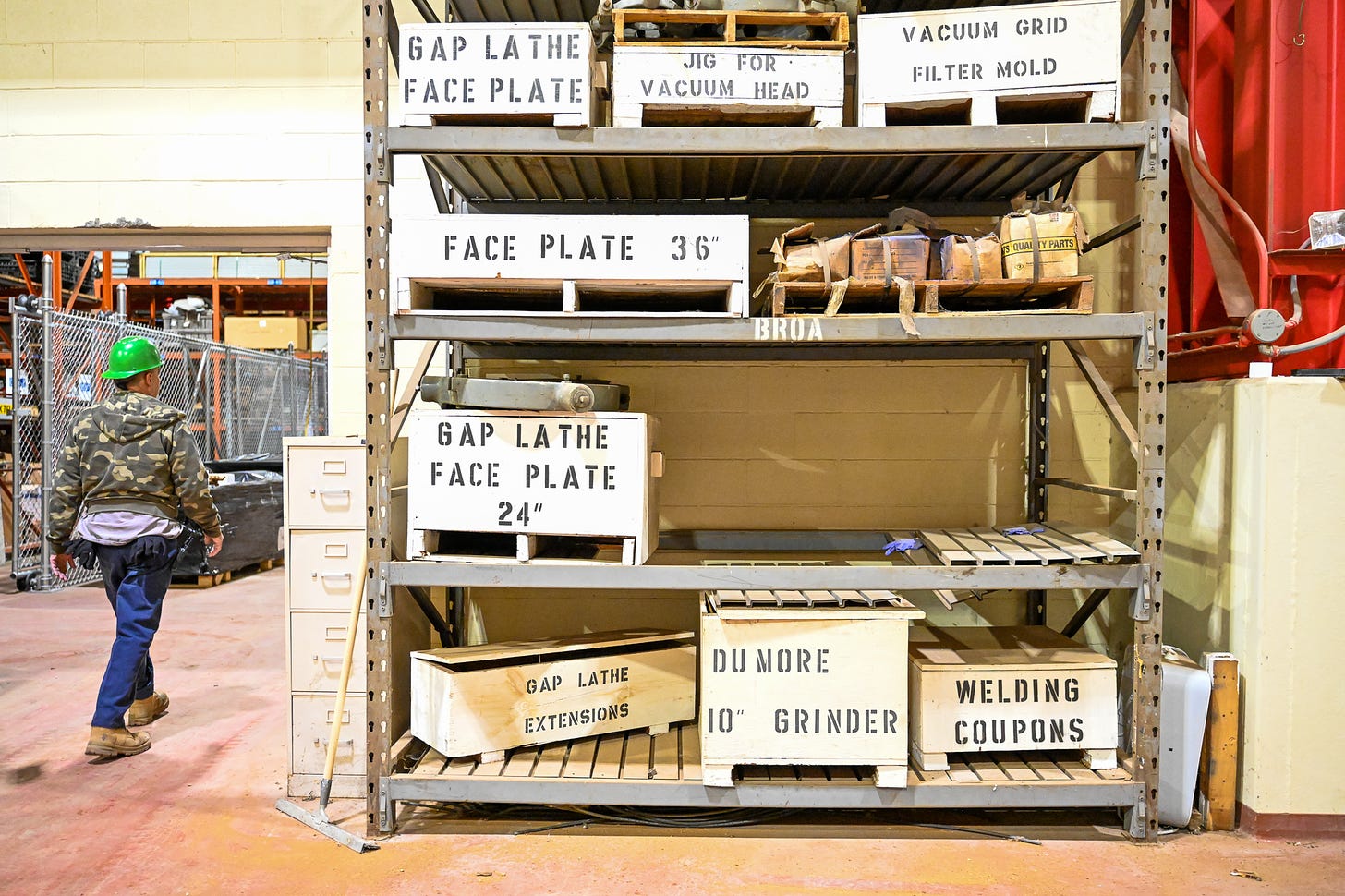 a tall wide shelving unit of boxes stretches from floor to ceiling. every shelf has beige crates labeled in black stencil-painted text. on the left a man walks forward past the shelves.