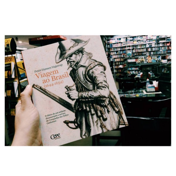 Livro “Viagem ao Brasil”, com gravura antiga de um soldado segurando um mosquete na capa. Ao fundo, estantes de uma livraria.