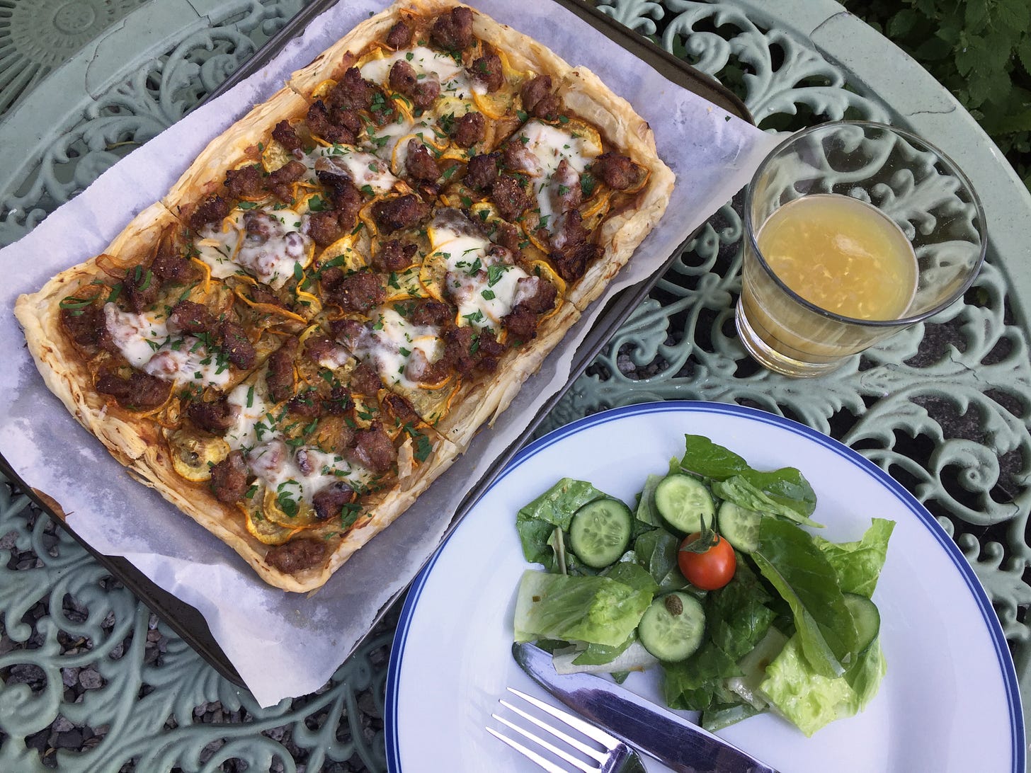 On a green wrought-iron table, a puff pastry tart with sausage, yellow zucchini, herbs, and patches of provolone sits on a parchment-lined baking sheet. To the right is a glass of IPA, and in the bottom corner is a plate with salad next to a knife and fork.