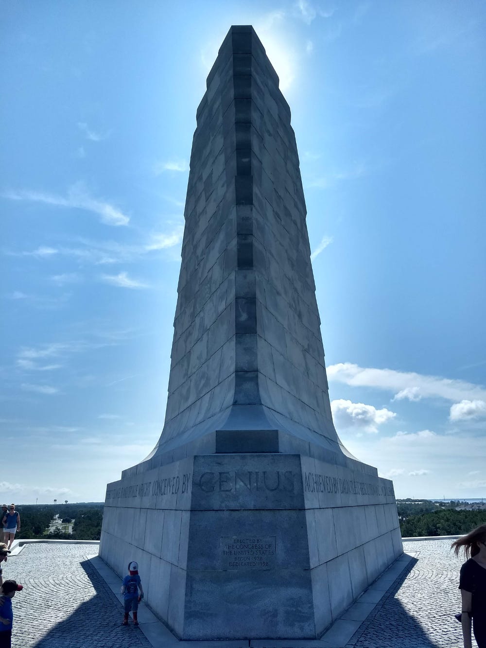 A contrasty photography of the Wright Brothers Memorial, with the sun behind it. The word "genius" is prominent on the front of the very tall, wedge-shaped sculpture.