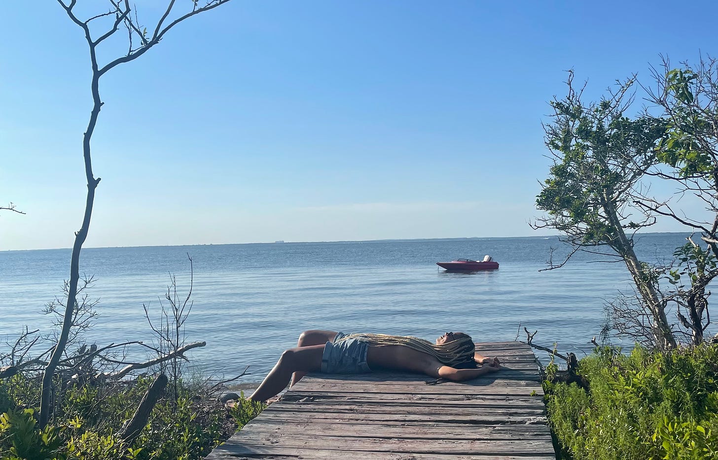 Me laying on a wooden dock surrounded by small trees, with a boat on the water in the distance.