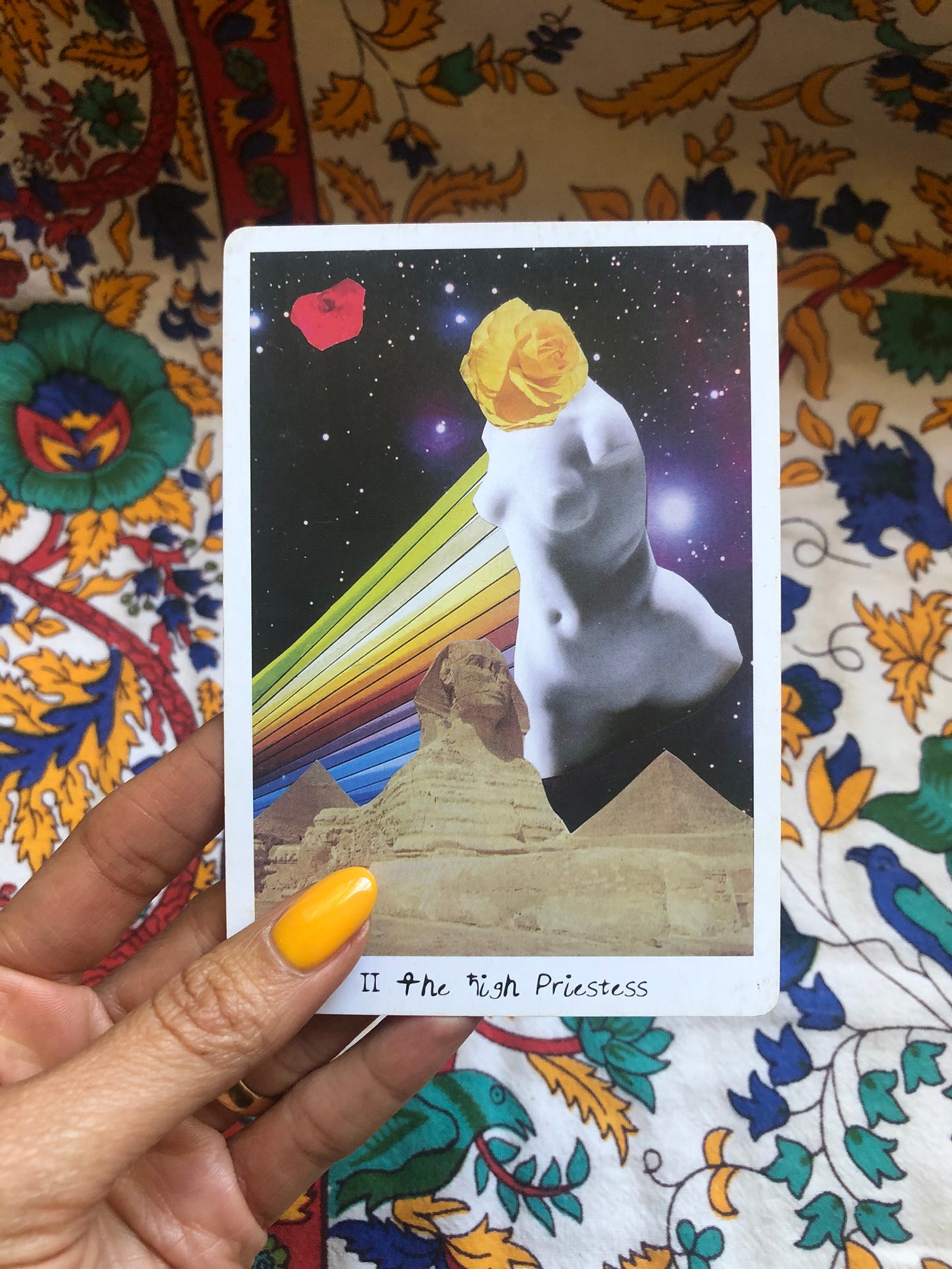 A hand with fingernails painted with yellow nail polish holds up The High Priestess tarot card from The Lioness Oracle Tarot deck. The card features a statue/bust of a female body, with a yellow rose for a head. There is a cut-out photo of the sphinx statue and pyramids in Egypt, and a rainbow featured on the card as well, against the backdrop of a starry night sky.